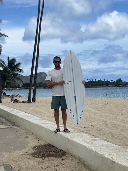 Harley Ingleby standing with the Harley Ingleby Mid 4 surfboard upright at Ala Moana Beach, Honolulu