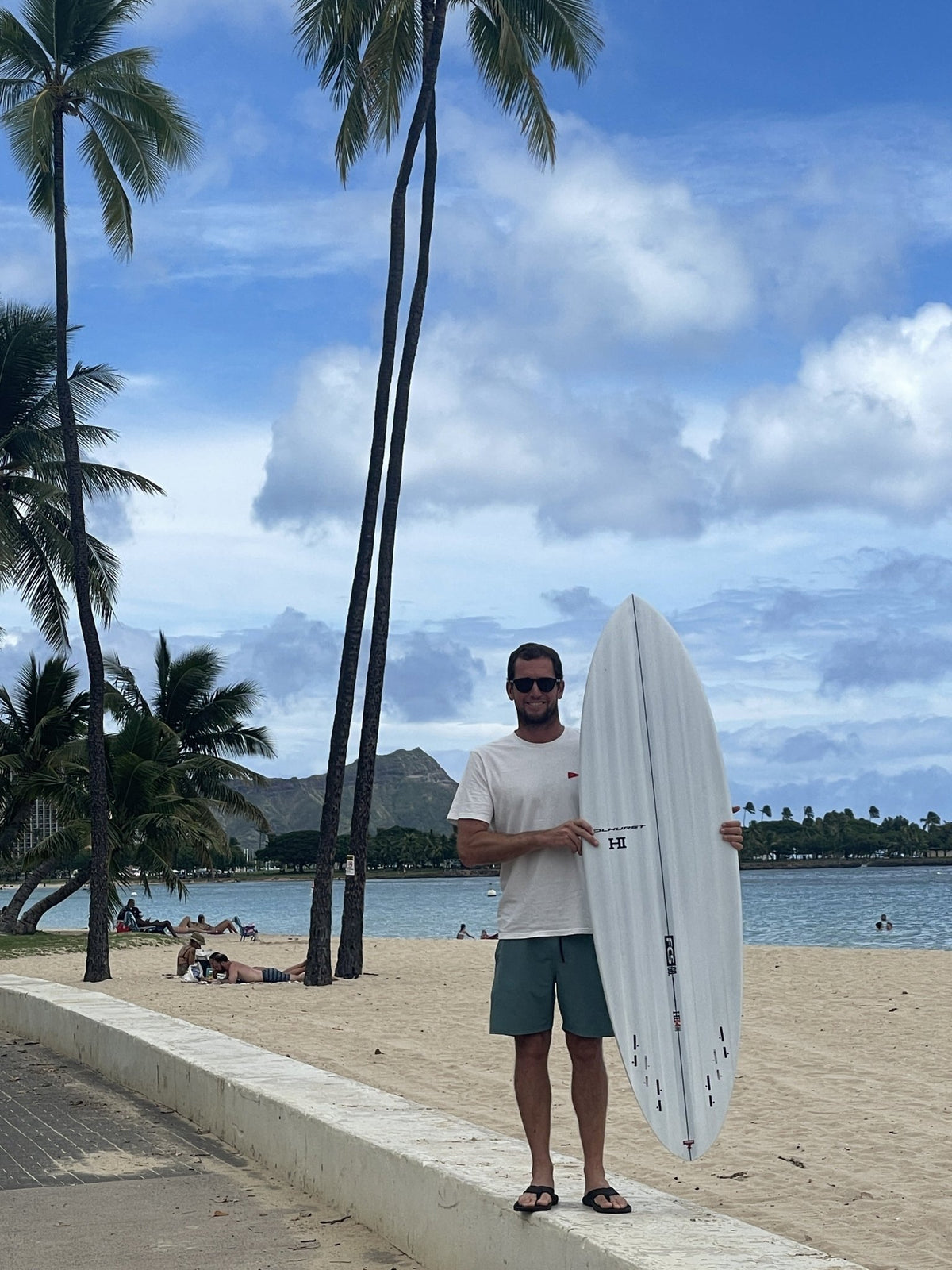 Harley Ingleby with the Harley Ingleby Mid 4 surfboard under palm trees at Ala Moana Beach, Honolulu