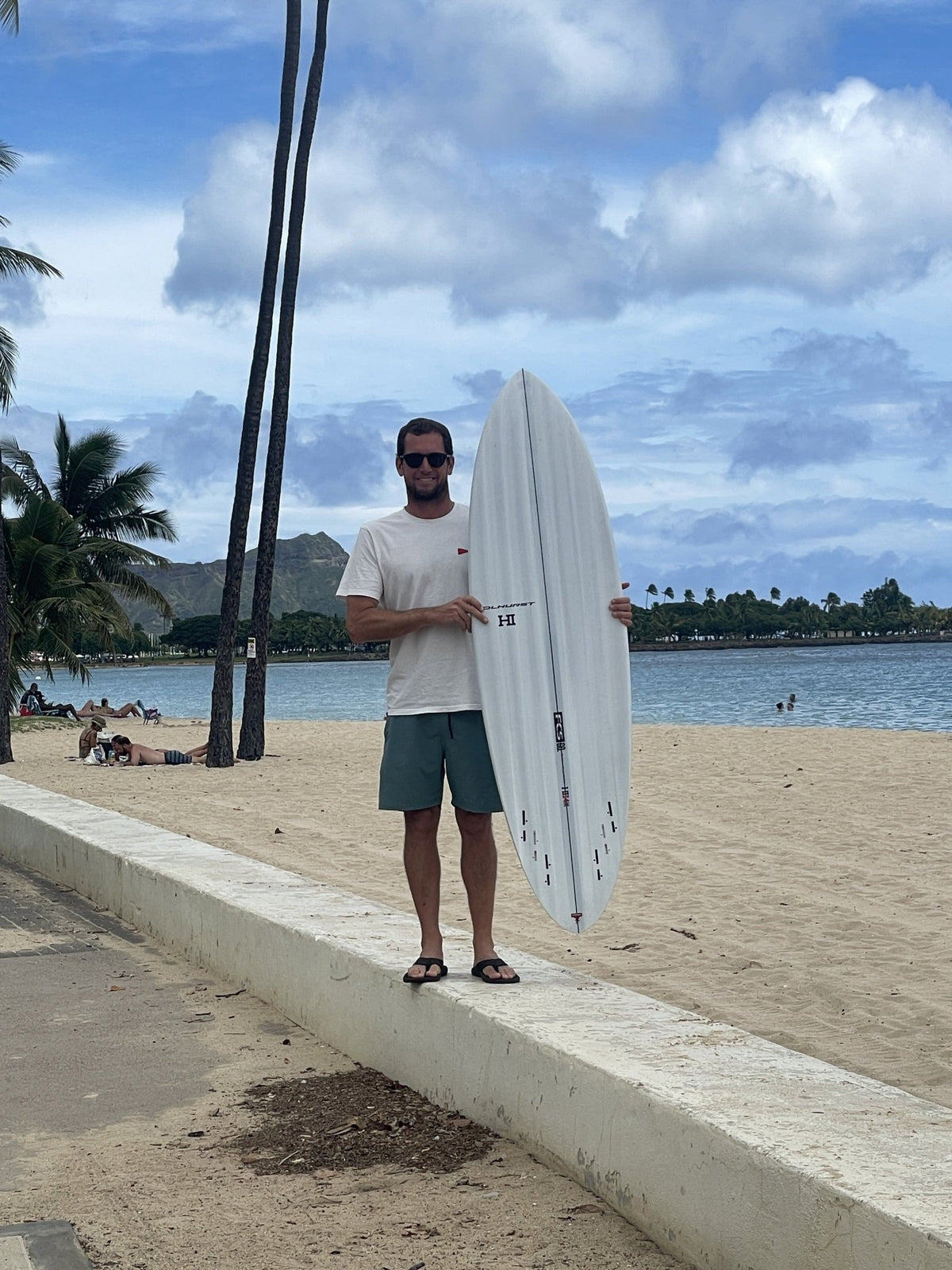 Harley Ingleby standing with the Harley Ingleby Mid 4 surfboard upright at Ala Moana Beach, Honolulu